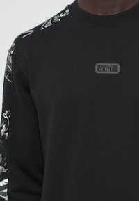 Sweatshirt preto com mangas de padrão floral, tecido texturizado e uma etiqueta retangular a dizer "VERSACE JEANS COUTURE."