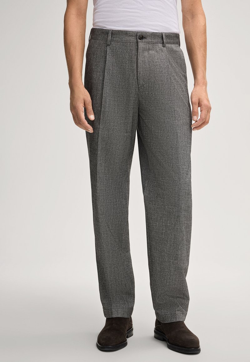 Uomo che indossa pantaloni eleganti grigi a quadri, camicia bianca infilata nella cintura e scarpe eleganti in suede marrone scuro, in piedi contro uno sfondo neutro.