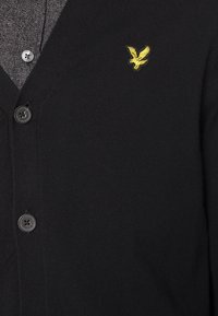 Schwarze Strickjacke aus gestricktem Material; hat einen V-Ausschnitt, vier Knöpfe und ein kleines gelbes Adlerlogo auf der linken Brust.