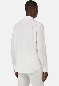 Camicia bianca di lino a maniche lunghe con orlo leggermente curvato, morbida al tatto, con polsini abbottonati e colletto semplice, vista dal retro.