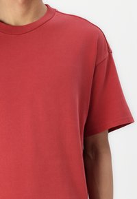 Tricou roșu din bumbac, cu guler rotund, mâneci scurte și o textură netedă. Prezintă o croială lejeră, cu cusături pe umeri.