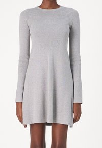 Donna che indossa un vestito a maniche lunghe di maglia grigio chiaro con un top aderente e una gonna svasata, caratterizzato da un sottile logo in rilievo sul petto.