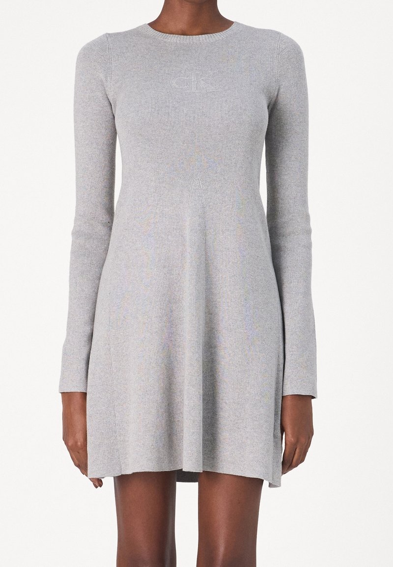 Donna che indossa un vestito a maniche lunghe di maglia grigio chiaro con un top aderente e una gonna svasata, caratterizzato da un sottile logo in rilievo sul petto.