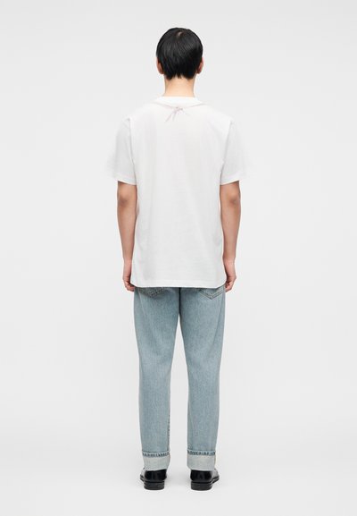 Άσπρο βαμβακερό t-shirt με άνετη εφαρμογή, με κοντά μανίκια και διακοσμητικό ροζ φιόγκο στο πίσω μέρος του λαιμού, συνδυασμένο με ανοιχτόχρωμο τζιν.