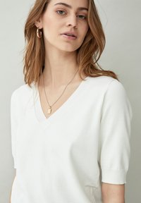 Suéter de punto blanco de manga corta con un diseño de cuello en V. Presenta una textura suave y sutiles acentos acanalados. Se lleva con un collar delicado.