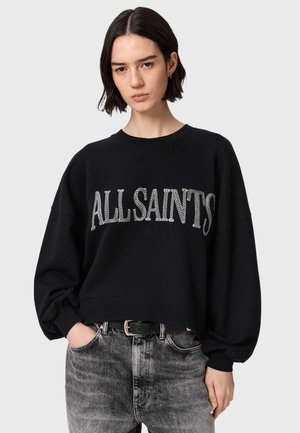 Kobieta o krótkich ciemnych włosach ubrana w czarną, oversize'ową bluzę z białym napisem "ALLSAINTS" oraz szare, wyblakłe dżinsy na szarym tle.