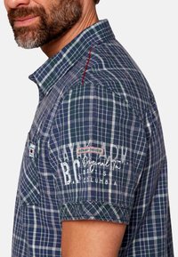 Camicia a maniche corte da uomo a quadri blu e verdi con una cucitura accentuata rossa. Presenta un logo e un design testuale sul lato sinistro del petto. Materiale in cotone.