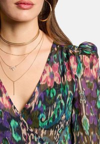 Robe plissée colorée avec un décolleté en V, ornée d'un mélange de motifs violets, verts et roses. Des colliers en or empilés et des boucles d'oreilles créoles viennent compléter le look.