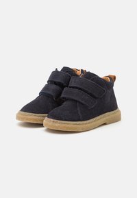 Botas de tobillo de ante azul marino con cierre de velcro, forro en color caramelo, suela de goma y detalle de costura a lo largo del borde. Diseñadas para ofrecer comodidad y soporte.