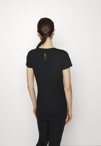 Kvinna med flätat hår som bär en åtsittande svart T-shirt och svarta leggings, stående mot en plain vit bakgrund och vänd bort.