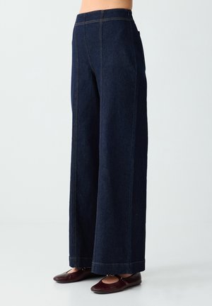 Dunkelblaue, weit geschnittene Jeanshose, getragen mit dunkelbraunen flachen Schuhen, von der Taille bis zu den Füßen vor hellem Hintergrund gezeigt.