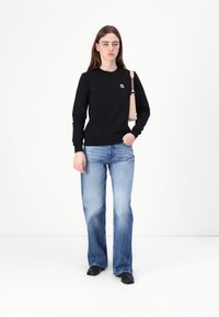 Felpa nera con un piccolo logo, abbinata a jeans chiari a zampa d'elefante. La modella tiene una borsa beige, in piedi su uno sfondo bianco.