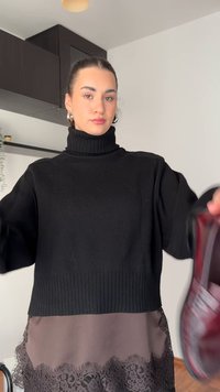 Černý nadměrně velký rolákový svetr s žebrovanými detaily, vrstvený přes hnědé šaty s krajkovým lemem. Drží se červený předmět.