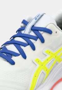 Baskets de course blanches avec des lacets bleus, un logo Asics jaune bien en vue et une tige texturée. La semelle est d'un rose vif.