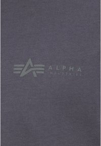 Camiseta de algodón gris con un logo impreso que muestra una "A" estilizada y el texto "ALPHA INDUSTRIES" en un color contrastante. Textura suave.