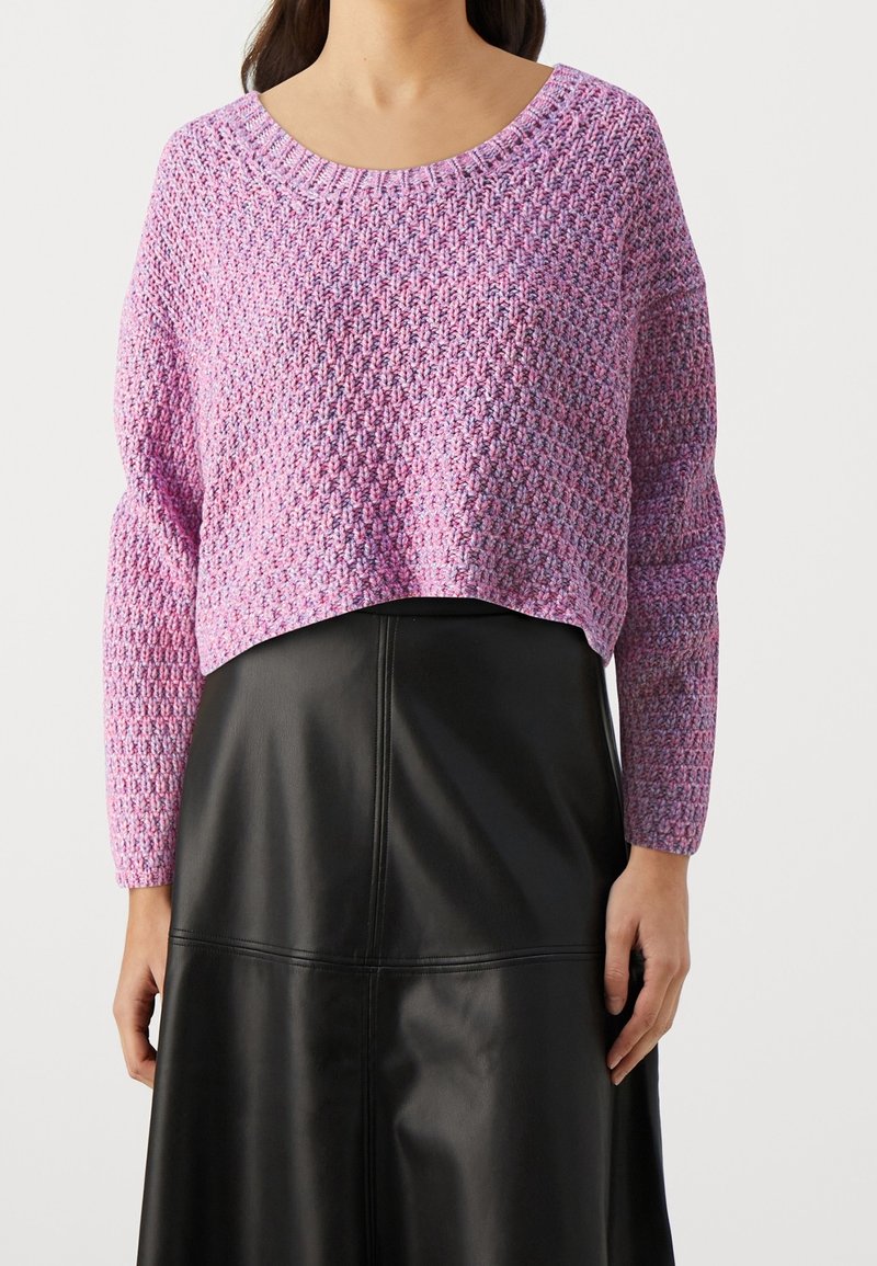 Femme portant un pull en maille violet texturé à manches longues et une jupe en cuir noire, debout devant un fond uni.
