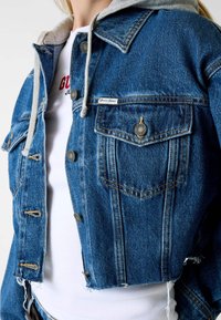 Przycięta kurtka jeansowa w kolorze niebieskim z szarą podszewką kaptura, wyposażona w metalowe guziki, przednie kieszenie i postrzępione wykończenia na dole.