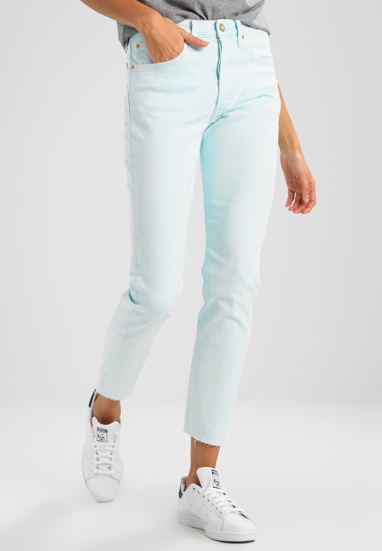 Jeans de denim azul claro con un corte ajustado, cinco bolsillos, costuras visibles y dobladillos deshilachados, combinados con zapatillas blancas y una camiseta gris.