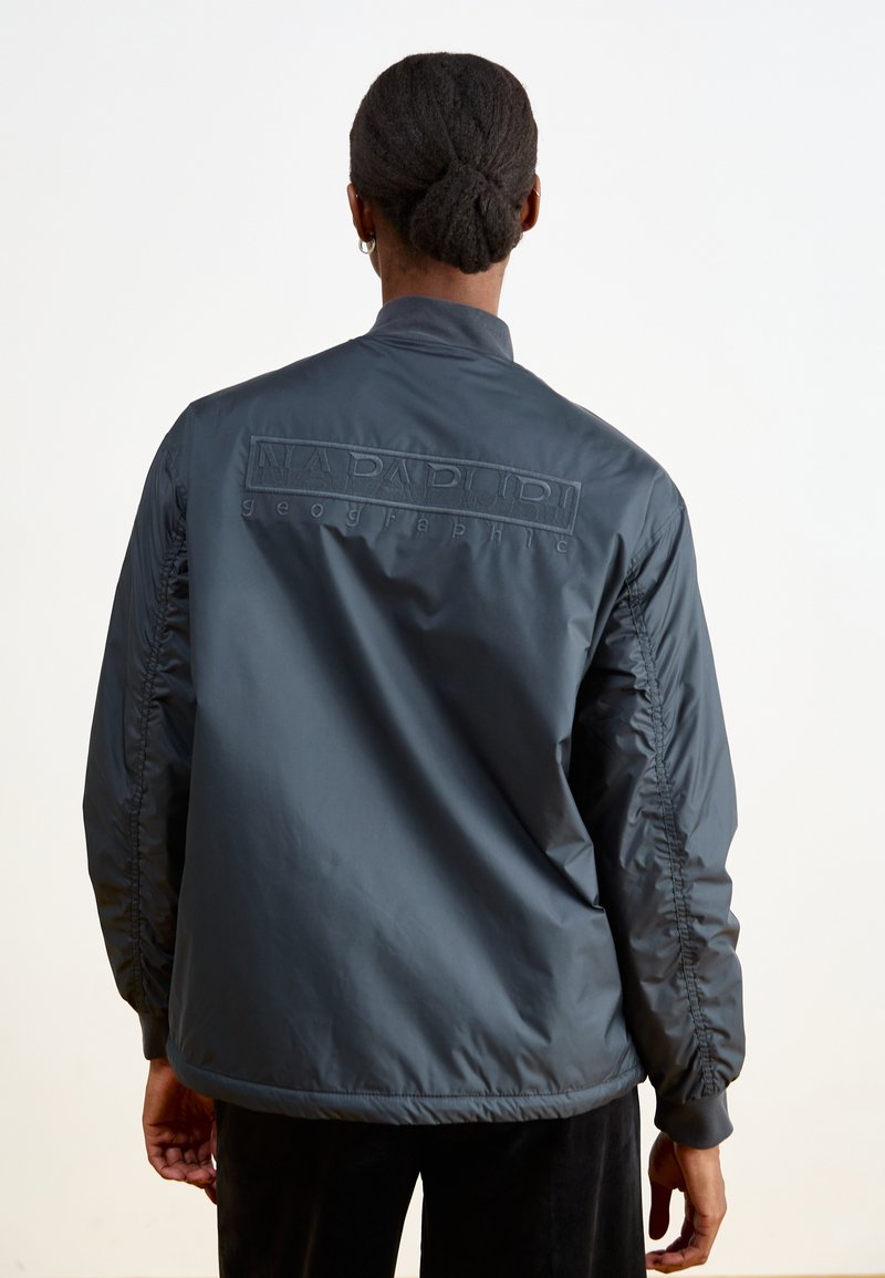 Ciemnoszara, ocieplana kurtka z elastycznymi mankietami, z teksturowanym logo na plecach. Materiał wydaje się gładki z delikatnym połyskiem.