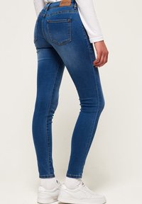 Jeans de mezclilla azul ajustados con una textura suave, cinco bolsillos y un parche de cuero en la cinturilla trasera; usados con zapatillas blancas.