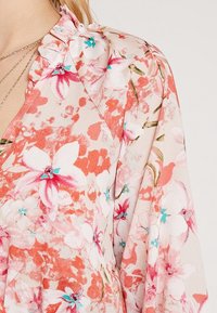 Tissu à imprimé floral dans des tons de rose et corail avec des touches de turquoise ; présente un col à volants et une texture douce et légère.