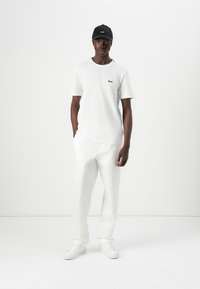 T-shirt en coton blanc avec le logo "BOSS" noir, associé à un pantalon blanc sur mesure et des baskets blanches. Le modèle porte une casquette noire.