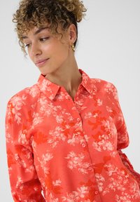 Frau mit lockigem Haar, die goldene Creolen trägt und ein korallenfarbiges, blumiges Hemd mit Knöpfen anhat, nach unten schaut und leicht lächelt.