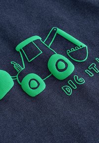 Dunkelblauer Stoff mit einem grünen grafischen Motiv eines Cartoon-Traktors mit kräftigen Konturen und dem Text "DIG IT!" in einer verspielten Schriftart.
