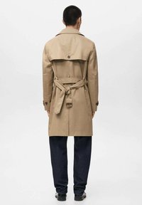 Beige trenchcoat met een ceintuur in de taille, schouderepauletten en knopenaccenten, gecombineerd met donkere jeans en zwarte schoenen. Zichtbare naden en textuur.