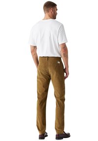 Pantaloni in velluto a coste marroni con vestibilità dritta, abbinati a una maglietta bianca semplice. Marchio visibile sulla tasca posteriore.