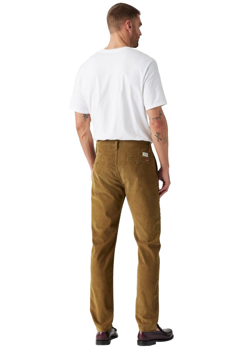 Pantalon en velours côtelé marron à coupe droite, associé à un t-shirt blanc uni. Marque visible sur la poche arrière.