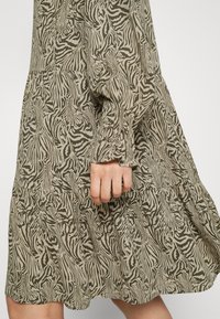 Abito a strati di colore verde oliva con stampa zebra nera, maniche lunghe e gonna arricciata, realizzato in tessuto leggero. Mano mostrata mentre tiene il tessuto.