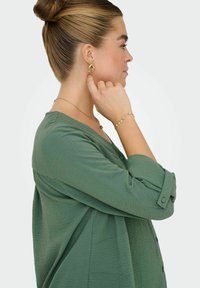 Boucles d'oreilles en or avec des accents texturés, un haut vert à boutons, et des bracelets en or superposés. Le mannequin est de profil avec les cheveux en chignon.