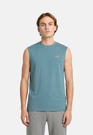 Giovane uomo che indossa una maglietta senza maniche turchese con un piccolo logo sul petto e pantaloni grigi, in piedi davanti a uno sfondo chiaro.