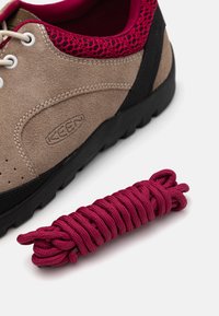 Sneaker en suède beige avec des accents noirs, un col en mesh rose, et des détails perforés, accompagnée d'un paquet de lacets bordeaux torsadés.