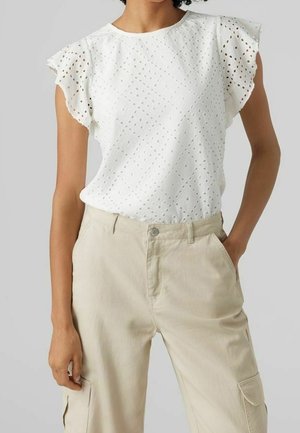 Witte blouse met korte mouwen van eyelet-stof en gerimpelde schouders, gecombineerd met beige cargobroek met hoge taille, één hand in de zak.