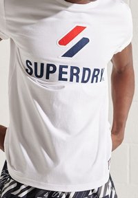 Bílý bavlněný tričko s velkým logem "SUPERDRY" v námořnické modré a červené barvě s geometrickým prvkem nahoře. Krátké rukávy, běžný střih.