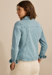 Chaqueta de mezclilla azul clara con un diseño ajustado, puños abotonados y yugo trasero. La tela tiene una textura suave y detalles de costura sutiles.