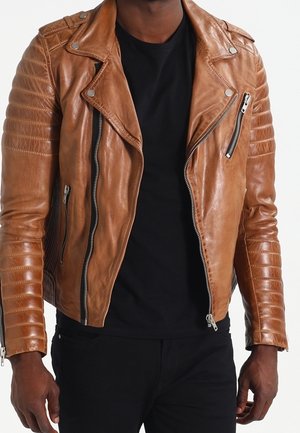Hombre con chaqueta de cuero marrón con mangas acolchadas y múltiples cremalleras plateadas sobre una camisa negra y pantalones negros.