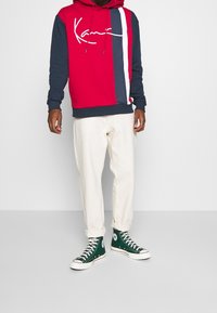 Hombre vestido con una sudadera roja y azul marino con rayas blancas y rojas, pantalones off-white remangados y zapatillas altas de color verde oscuro, de pie contra un fondo blanco.