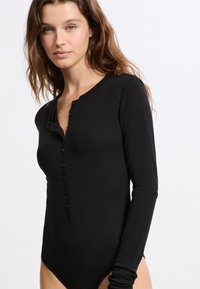 Body noir à manches longues en tissu doux avec un col rond et une fermeture à boutons sur le devant. Coupe ajustée mettant en valeur la silhouette.