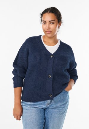 Navy knitted cardigan with a V-neck, button front, and oversized sleeves. Soft texture and relaxed fit, paired with light blue jeans.