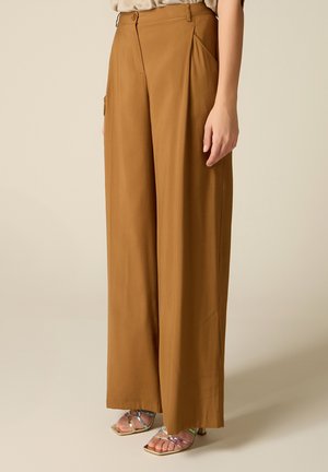 Mujer que lleva pantalones marrones de talle alto y pierna ancha combinados con sandalias de tiras abiertas con tacones medios sobre un fondo liso.