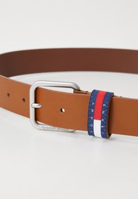 Cinturón de cuero marrón con una hebilla de metal plateada, que presenta una lengüeta azul con rayas rojas y blancas y el logo "TOMMY JEANS".