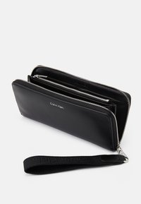 Musta nahkatalous, jossa on suuri vetoketju, useita lokeroita, tyylikäs suorakulmainen muoto ja kangasranneke, jossa on logo.