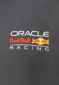 Γκρι ύφασμα με κεντημένο κείμενο: «ORACLE» σε λευκό, «Red Bull» σε κόκκινο και «RACING» σε λευκό, με διακοσμητικό λογότυπο Red Bull σε κίτρινο.