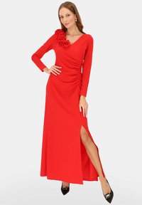 Czerwona sukienka z długim rękawem, z marszczeniami, dekoltem w kształcie litery V oraz kwiatowym akcentem na jednym ramieniu. Wysoki rozporek po lewej stronie.