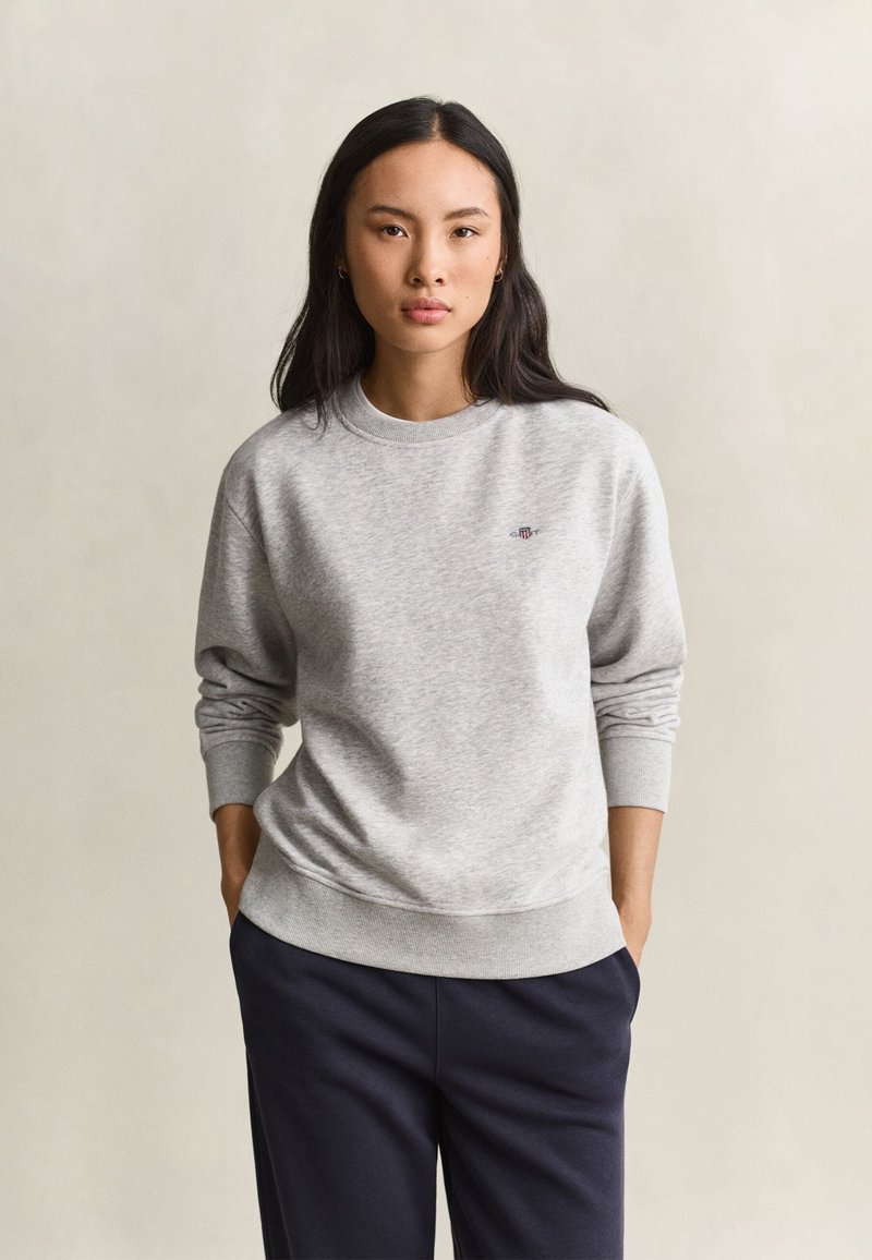 Femme aux longs cheveux foncés portant un sweat-shirt gris clair et un pantalon foncé, debout devant un fond beige uni.