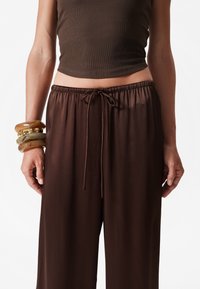 Brown wide-leg pants with an elastic waistband and drawstring detail, made of smooth material. Worn with stacked golden-brown bangles.