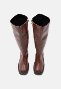Καφέ δερμάτινες μπότες μέχρι το γόνατο με λείο φινίρισμα, τετράγωνη μύτη και ραφές σε αντίθεση. Διαθέτουν φερμουάρ στην εσωτερική πλευρά.